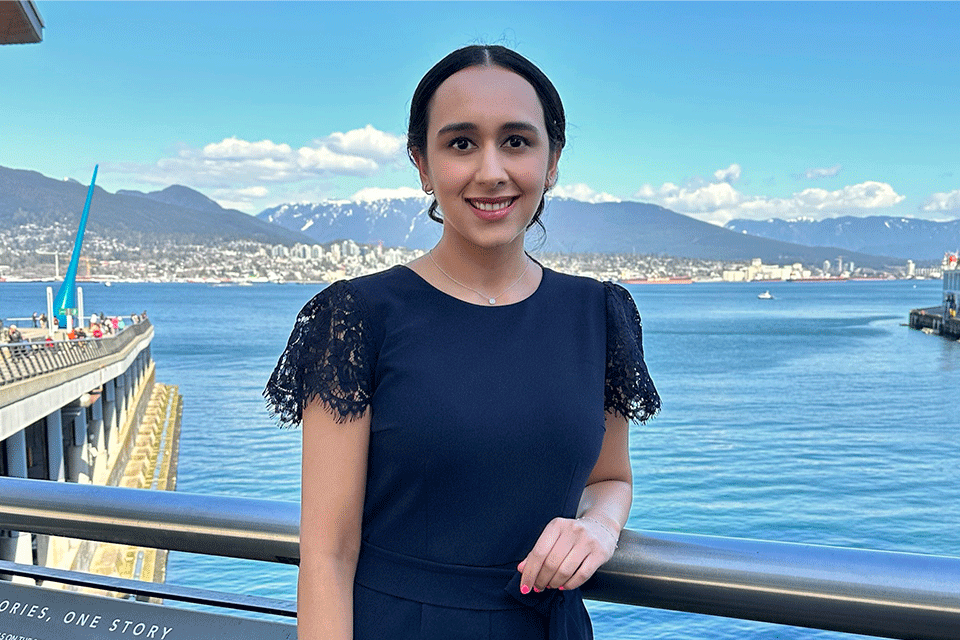Photo of woman (Rising Star Amanpreet Powar) wearing a navy blue top and posing against a grey railing with the sea and Vancouver mountains in the background