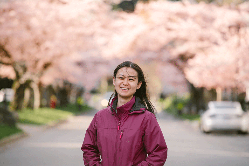 Photo of woman (Rising Star Amelia Dai) wearing a navy dark pink-purple top, smiling with cherry blossom trees in the background