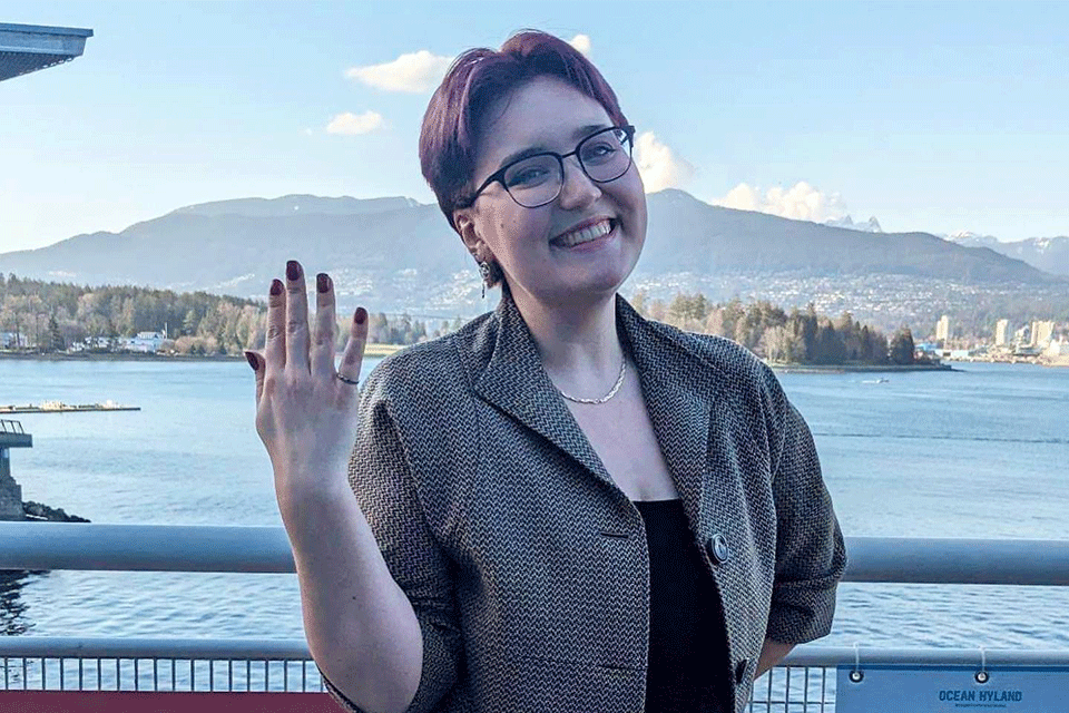 Photo of Rising Star Shannon Smyth wearing glasses and smiling showing off the palm of their hand whilst posing against a background of the sea and Vancouver mountains.ver mountains in the background