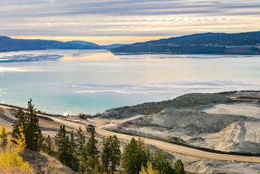 A large body of water, with mountains and sky in the background, and a mine access road and work trucks in the foreground.