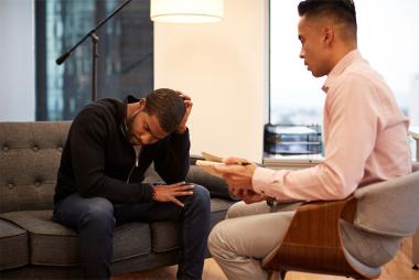 Two men sitting opposite each other in a therapy session, with client on a grey sofa looking down.
