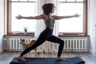 Woman stretching in a standing position with arms out whilst standing on an exercise mat. There is a dog behind her.