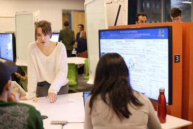 Student stands in front of a digital screen with a presentation on it, while speaking to two people across a table.