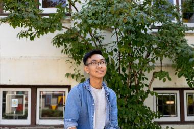 UBC School of Community and Regional Planning masters student William Canero smiles for the camera while gaxing upward and to the right. He is centered with a building and greenery behind him.