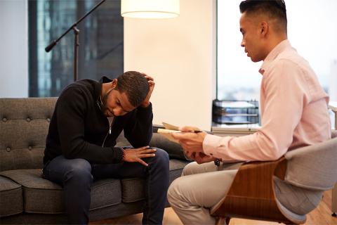 Two men sitting opposite each other in a therapy session, with client on a grey sofa looking down.