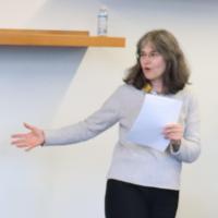 Woman in grey sweater, holding notes in one hand and gesturing off-screen with the other